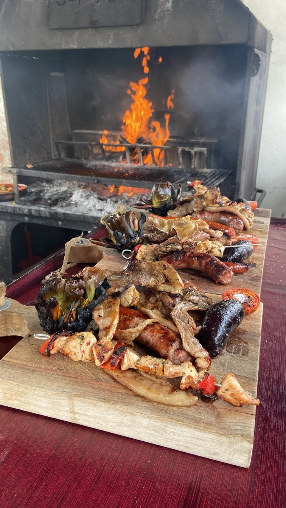 Parrillada de carnes y verduras sobre una tabla de madera, delante de la brasa encendida
